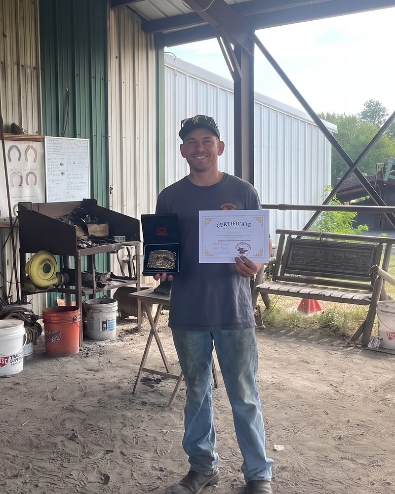 Luke Ure standing in a farrier's forge holding his certificate